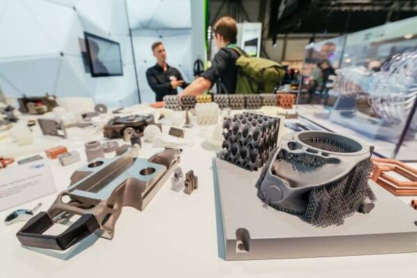 Impressionen von der Messe, zwei Personen im Hintergrund, im Vordergrund mehrere 3D-gedruckte Teile