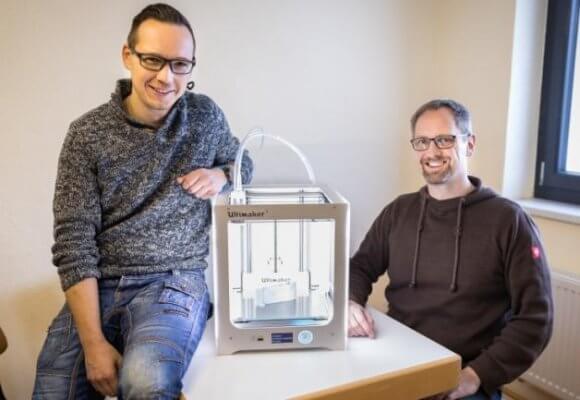 Andreas Kramer und Marcel Bochmann bei einem 3D-Drucker sitzend