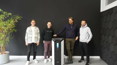 Vertreter von ASM und DyeMansion präsentieren die VX1 Glättanlage als Teil ihrer strategischen Partnerschaft im Bereich Vapor Smoothing. Gruppenfoto der Vertreter von ASM und DyeMansion mit der VX1 Glättanlage, die im Rahmen ihrer Partnerschaft für Vapor Smoothing im 3D-Druck vorgestellt wird.