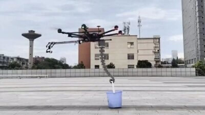 SpiRobs 3D-gedruckter Robotergreifarm hebt einen Eimer mit einer Drohne in einer Demonstration an 3D-gedruckter SpiRobs-Greifarm, der mit einer Drohne kombiniert einen Eimer hebt, vor städtischem Hintergrund