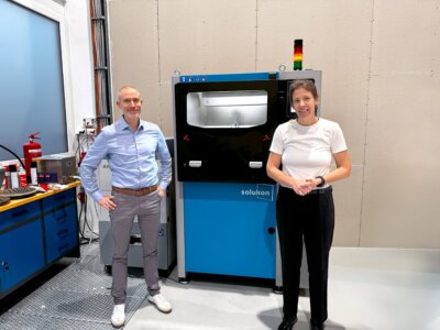 Andreas Hartmann und Hélène Huby vor der Solukon SFM-AT350-E für 3D-Druck-Nachbearbeitung. CEO von Solukon und CEO der Exploration Company vor der Solukon SFM-AT350-E, einer Anlage zur nachhaltigen Nachbearbeitung 3D-gedruckter Bauteile.
