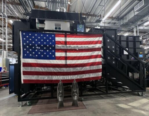 Sapphire XC 3D-Drucker von Velo3D in einer Produktionshalle mit amerikanischer Flagge und 3D-gedruckten Metallbauteilen