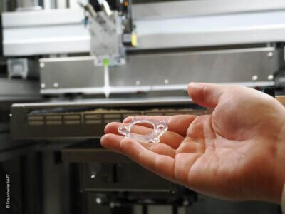 3D-gedruckte Silikondichtung aus der Studie des Fraunhofer IAPT zur additiven Fertigung in der Dichtungstechnik Nahaufnahme einer 3D-gedruckten Silikondichtung, gehalten von einer Hand, mit einem 3D-Drucker im Hintergrund