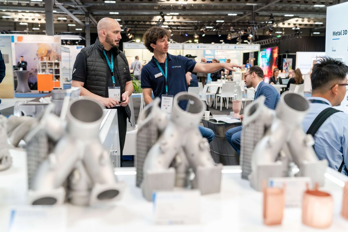 Besucher auf der rapid.tech 3D betrachten 3D-gedruckte Metallkomponenten an einem Messestand
