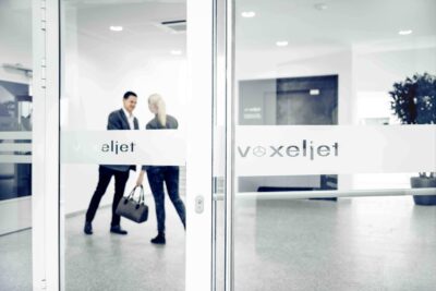 Eingangsbereich des voxeljet AG Hauptsitzes nach Ankündigung des Restrukturierungsverfahrens Zwei Personen im Eingangsbereich der voxeljet AG mit Firmenlogo auf Glastür, Symbolbild zur Restrukturierung