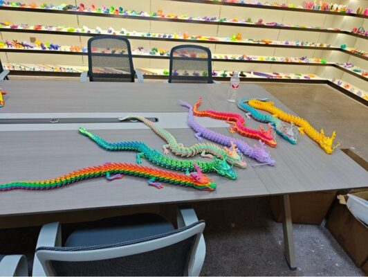 Mehrere farbige Drachenmodelle aus dem 3D-Druck liegen auf einem Konferenztisch, gedruckt mit FLSUN T1 MAX Druckern im Praxistest