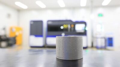 Zylindrischer, metallischer 3D-Druck-Ventiltrim von IMI vor einem industriellen 3D-Drucker